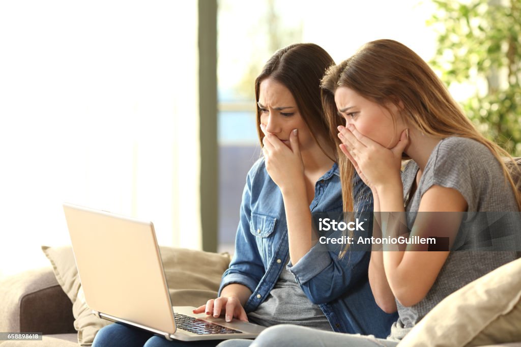 Worried roommates on line with a laptop Two worried roommates reading bad news or inappropriate content on line with a pc sitting on a couch in the living room in a house interior Identity Stock Photo