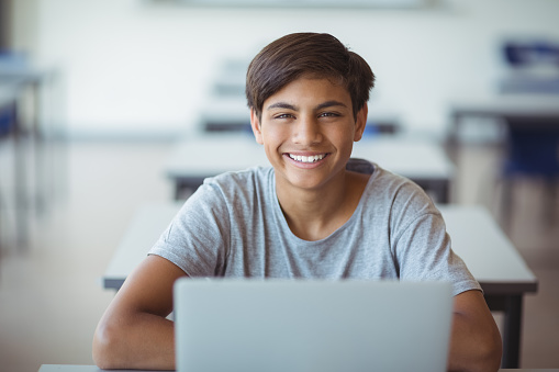 Portrait of happy schoolboy sitting with laptop in classroom at school