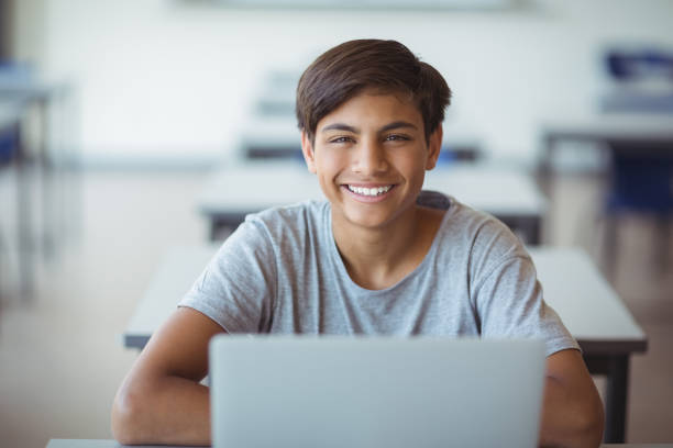 Portrait of happy schoolboy sitting with laptop in classroom Portrait of happy schoolboy sitting with laptop in classroom at school teenage boys stock pictures, royalty-free photos & images