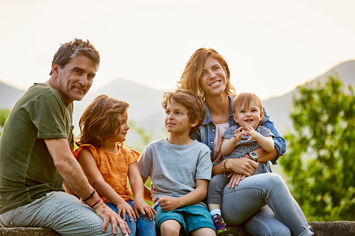 Portrait of happy parents with children. Family is sitting on retaining wall against sky. They are in casuals.