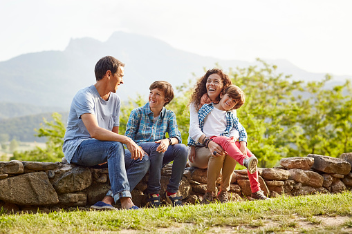 Happy family sitting on stone wall. Smiling parents and children are talking. They are spending leisure time on sunny day.