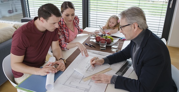 Architect explaining blueprint to couple in their home, daughter drawing at the table.