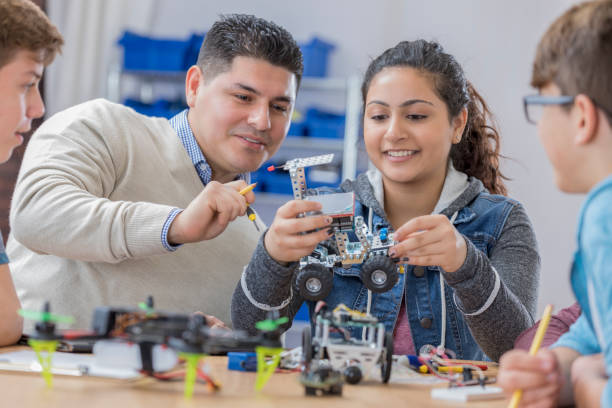 Helpful teacher works with students in technology class Confident mid adult Hispanic teacher helps a female student with a robotics assignment. The student is holding a robotic vehicle. Robot parts are on the table. high school teacher stock pictures, royalty-free photos & images