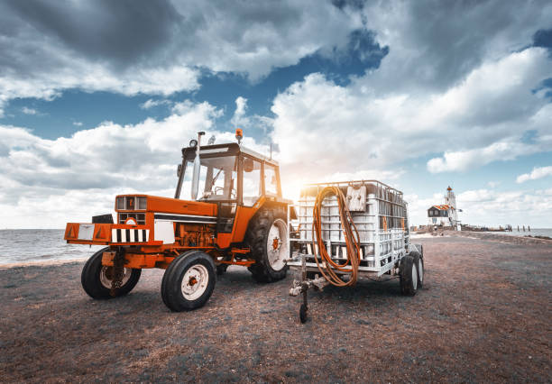 red tractor with trailer against lighthouse and blue overcast sky in spring at sunset. agricultural tractor. agricultural machinery and machines in bright sunny day. landscape with vintage toning - agricultural machinery retro revival summer farm imagens e fotografias de stock