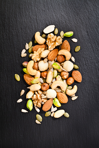 Vertical shot of mixed nuts arranged in a heap at the center of a black slate table. Nuts included in the composition are pistachios, hazelnut, pine nut, almonds, pumpkin seeds, macadamia nuts, sunflower seeds, peanuts, cashew and walnuts. Predominant color is brown and gray. DSRL studio photo taken with Canon EOS 5D Mk II and Canon EF 100mm f/2.8L Macro IS USM