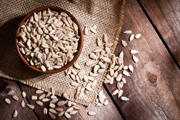 Top view of a brown bowl filled with organic sunflower seeds shot on rustic wood table. Some almonds are out of the bowl on a burlap. Predominant color is brown. DSRL studio photo taken with Canon EOS 5D Mk II and Canon EF 100mm f/2.8L Macro IS USM