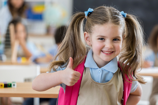 Young kindergartener is excited about the first day of school. She is giving a thumbs up. She is wearing a backpack and school uniform.