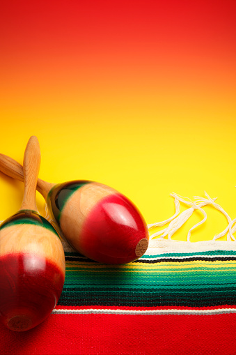 A still life of a pair of maracas on a multi-colored Mexican blanket that sits on a background that gradates from bright yellow to orange.