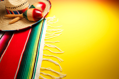A still life of a pair of maracas on a sombrero and a multi-colored Mexican blanket that sits on a background that gradates from bright yellow to orange.
