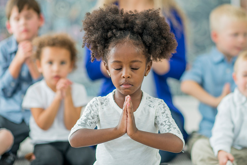 Young girl sits with her eyes closed and palms together praying while in front of a Sunday school class consisting of a teacher and young children