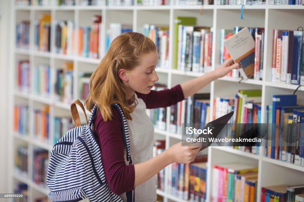 Schoolgirl using digital tablet while selecting book in library Schoolgirl using digital tablet while selecting book in library at school Library Stock Photo