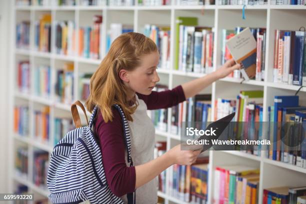 Schoolgirl Using Digital Tablet While Selecting Book In Library Stock Photo - Download Image Now