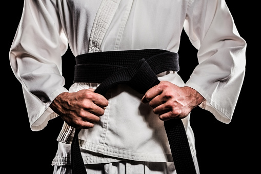 Man in keikogi tying black belt on dark background, closeup. Martial arts uniform