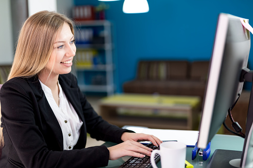 young, attractive businesswoman sitting at her desk with computer monitor in modern office, smiling happily.