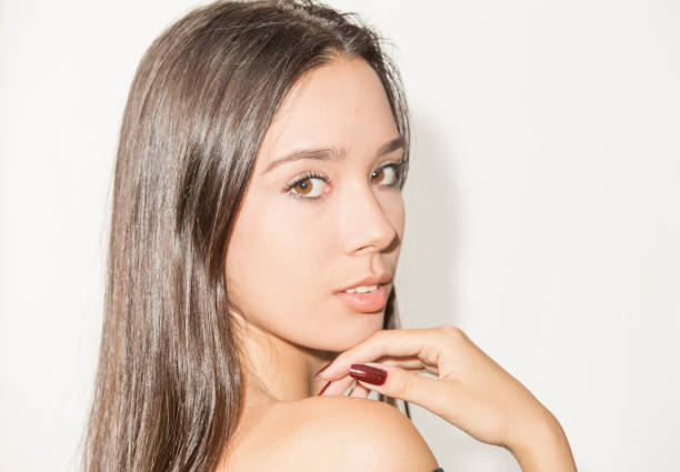Young lady poses for this close-up portrait. She is turning her head to the side where the camera is to look over her right shoulder where she put her right hand. She is latin, with brown eyes and hair. Her hair is brown, long and smooth. She looks like in her twenties.