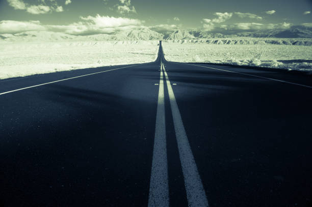 A freshly paved road cuts through the California desert. Horizontal, toned black and white image shot with an infrared camera for high contrast. Low angle shot from the center of the road.