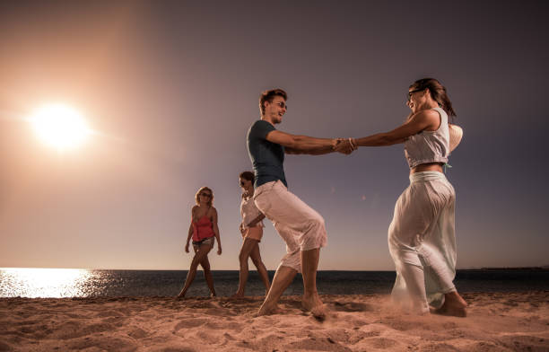 Young happy couple having fun on the beach while spinning. Two women are walking in the background.