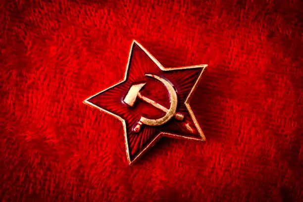Old Soviet badge with the red star, a sickle and a hammer reminiscent of the cold war era worn by the soldiers of the red army on their hats, isolated on red velvet with a grungy aesthetic