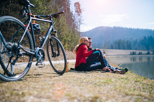 Sports couple is having a break during cycling. They are sitting on the floor and observing. Sun is shining and nature is around them.