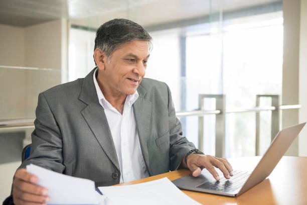 Senior businessman using laptop and looking at screen he is holding a document in one hand and typing on the keyboard with the other sri lanka business stock pictures, royalty-free photos & images