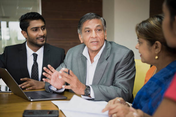 Serious senior businessman with laptop talking in meeting Man explaining to colleagues in meeting room, young man listening and smiling, woman watching as senior man makes hand gestures sri lanka business stock pictures, royalty-free photos & images
