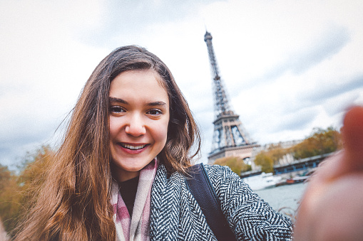 Young beautiful girl holding smart phone and taking selfie pictures. She is happy, smiling enjoying and looking at her camera at coastline of Seine river against Tour Eiffel in Paris, France.