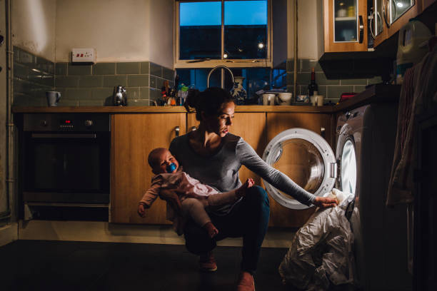 Multi-Tasking Mother Young mother is holding her crying baby daughter on her hip while trying to load the washing machine. working hard stock pictures, royalty-free photos & images