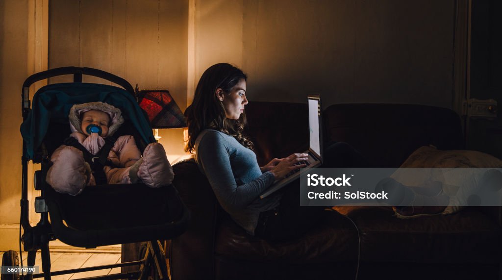 Multi-Tasking Mother Young mother is working on a laptop on the sofa in her home with her baby daughter sleeping in the pushchair next to her. Working Mother Stock Photo