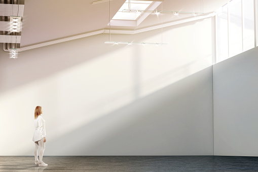 Woman walking near blank white wall mockup in modern gallery with sunset. Girl admires a clear big stand mock up in museum with contemporary art exhibitions. Large hall interior, sunrise from windows