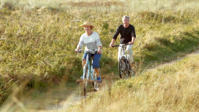 Mature couple bike riding on path along sunny beach grass