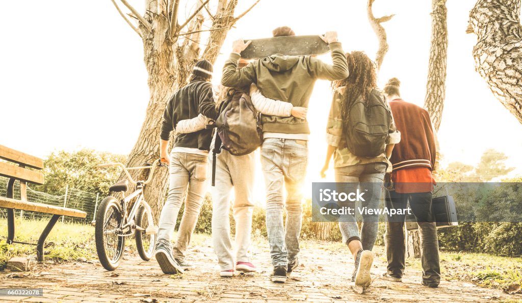Group of urban friends walking in city skate park with backlighting at sunset - Youth and friendship concept with multiracial young people having fun together - Warm retro filter with soft focus Teenager Stock Photo