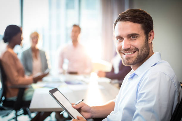 Man holding digital tablet while coworker interacting in the background Portrait of man holding digital tablet while coworker interacting in the background in the office sales executive stock pictures, royalty-free photos & images