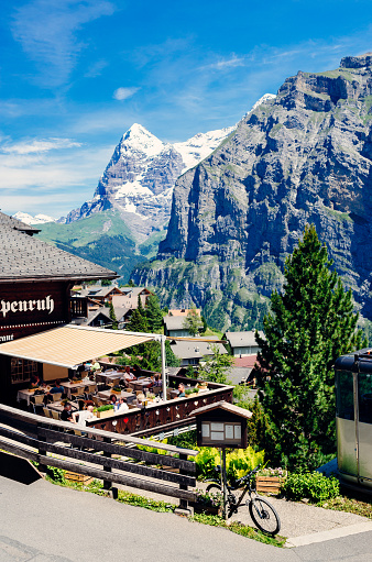 Tourists dining at the restaurant of the Hotel Alpenruh on Hinter der Egg in the Bernese Oberland alpine village of Murren, Switzerland, on a sunny summers day. The prominent steep snowy mountain on the skyline is the Eiger, with its famous North Face in profile.
