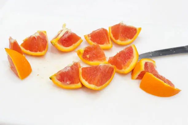 One bright orange fruit cut into twelve pieces with a knife on a white plastic cutting board close-up. An orange sliced into many chunks on a cutting board.