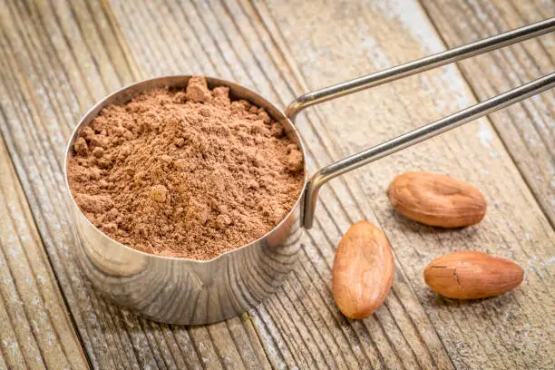 cacao powder in a metal measuring scoop against grunge wood background with some raw beans