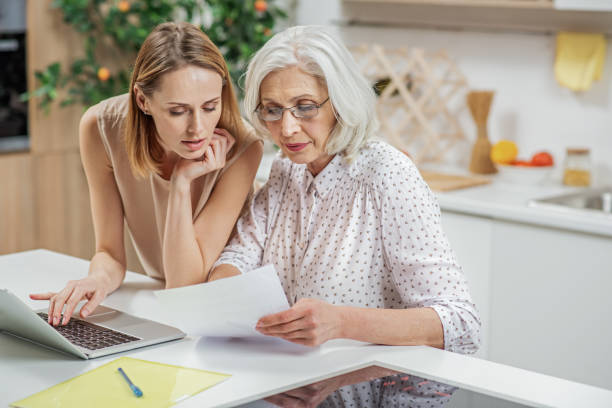 Friendly girl explaining some documentation to parent Senior woman is reading document with concentration. Her daughter is standing and helping her. She is typing on laptop convenience photos stock pictures, royalty-free photos & images