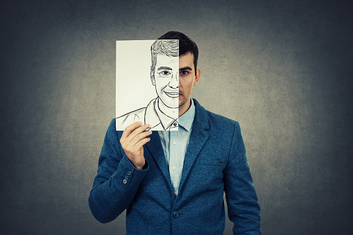 Portrait of a sad businessman hiding half his face using a white paper drawn with a fake happy emotion. Mask for hiding the real face expression, create new identity, grey wall background.