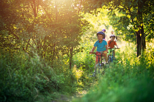 Outdoor portrait of a cheerful young boy in casual wear and protective blue helmet riding a balance bike along a leaf-strewn forest trail in dappled sunlight.