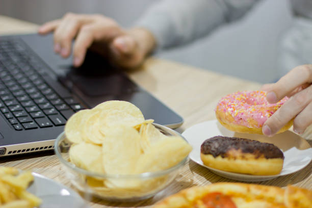 un homme travaille à un ordinateur et mange des fast-food. malbouffe : burger, sauce, pommes de terre, beignets, frites. - computer chip photos et images de collection