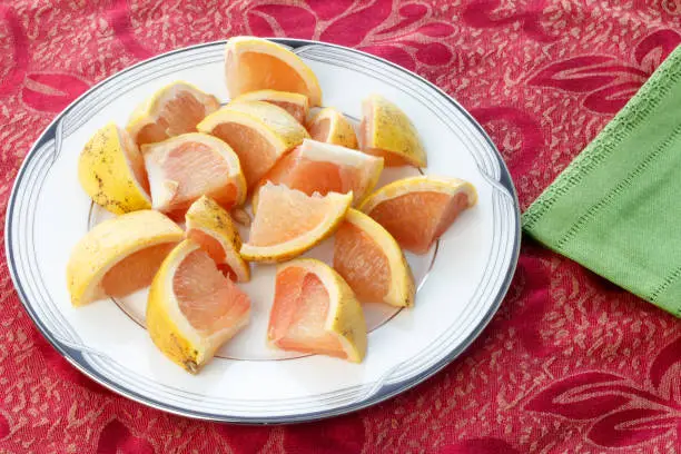 Many pieces of red grapefruit on a white round plate placed on a red placemat near a green cloth napkin. Red grapefruit cut into pieces on a dish ready to eat.