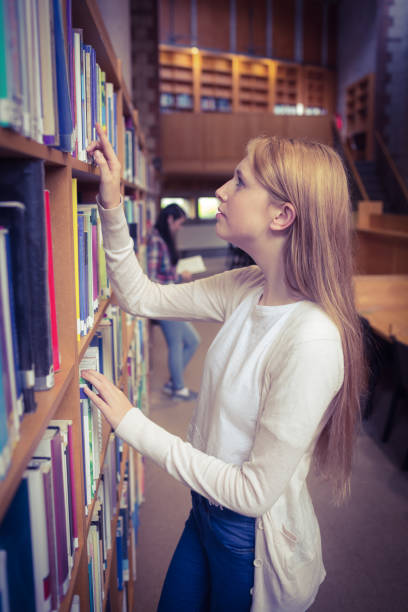 Thoughtful teenage girl reading books by shelves in library