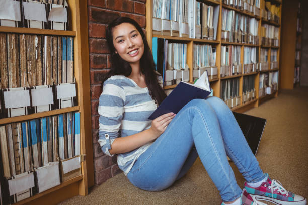 istock Smiling student sitting on the floor against wall in library reading book 660576056