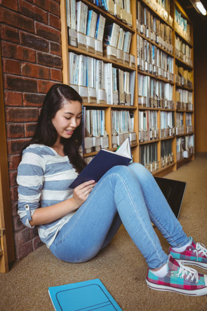 istock Smiling student sitting on the floor against wall in library reading book 660576092