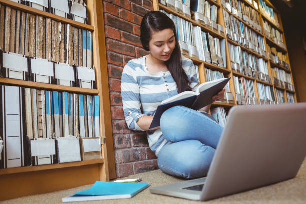 istock Smiling student sitting on the floor against wall in library studying with laptop and books 660576280