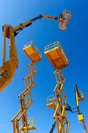 Scissor lift platform, cherry picker, aerial platform with bucket, articulating boom and other yellow construction cranes and machines, heavy industry, blue sky on background, bottom view