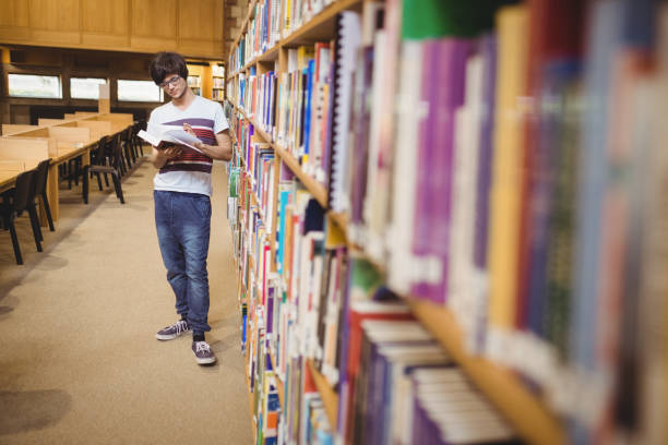 istock Young student reading book while standing near bookshelf 660707758
