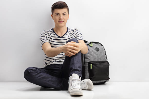 Teenager sitting on the floor next to a backpack and leaning against a white wall
