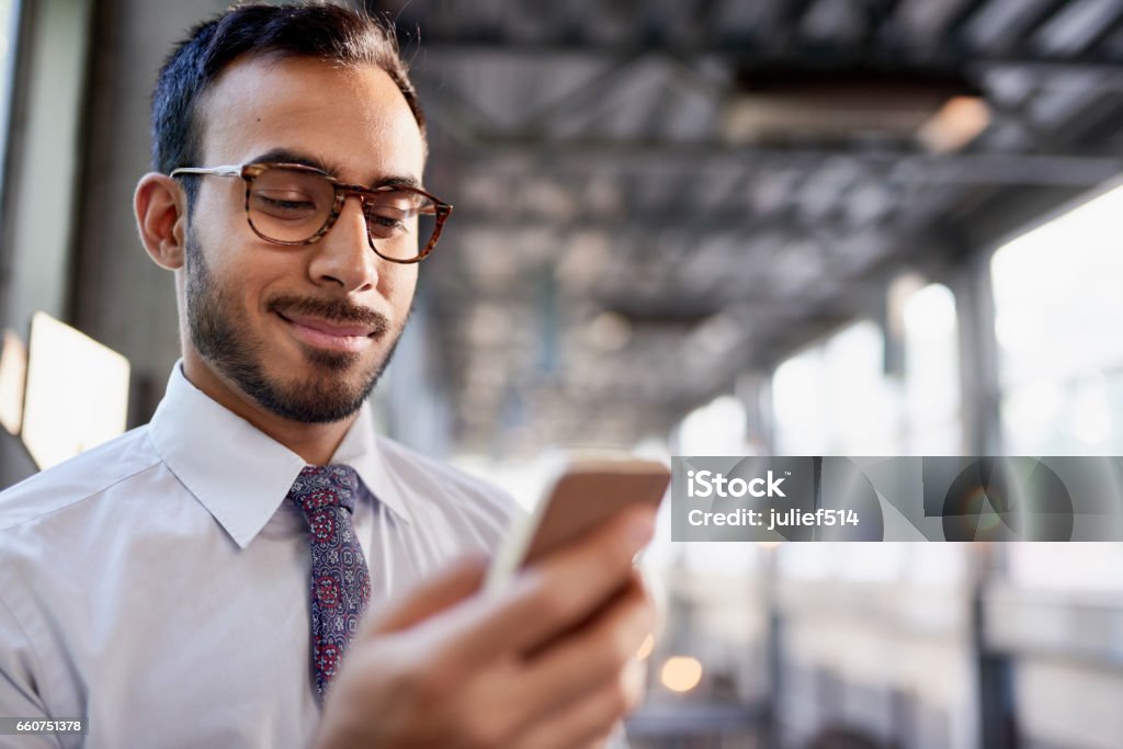 Indian businessman smiling confidently and surfing the net on a smartphone Portrait of a confident, happy business entrepreneur wearing fashionable tie and eyewear on his way to his next consultant meeting on the train platform Culture of India Stock Photo