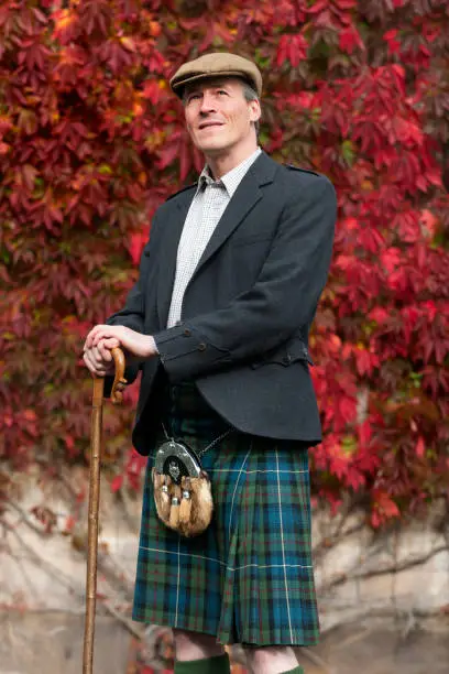 Elegant Scotchman in a traditional kilt and cloth cap posing with a walking stick or crook in front of a colorful red autumn creeper on a wall