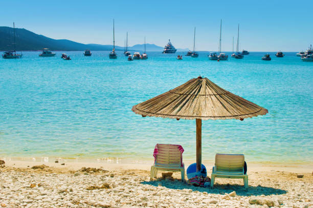 One yellow straw umbrella and two chairs on the pebble sandy beach One yellow straw umbrella and two chairs on the pebble sandy beach. Many yachts in the azure blue sea lagoon in a bay near the coast of an island. Blue cloudless sky. Dugi otok, Croatia dugi otok island stock pictures, royalty-free photos & images
